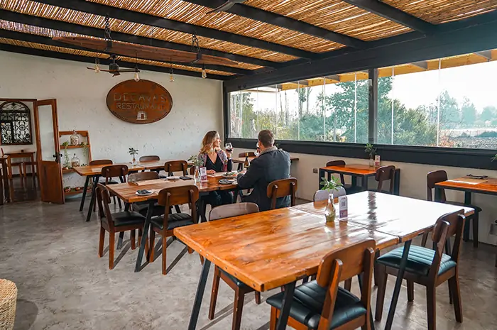 Restaurante interior “Decavas” en Hotel Santa Cruz con mesas de madera, piso de concreto, techo de listones y ventana con vista vegetal. Dos personas conversan en ambiente cálido y rústico. La escena transmite cotidianidad pausada, estética territorial y conexión con Cava Colchagua.