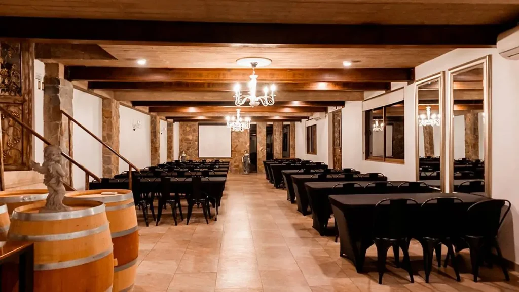 Salón interior en Hotel Santa Cruz con vigas de madera, muros de piedra, mesas vestidas en negro, barricas decorativas y luz cálida de araña. La escena transmite solemnidad vinícola, estética institucional y conexión con Cava Colchagua.