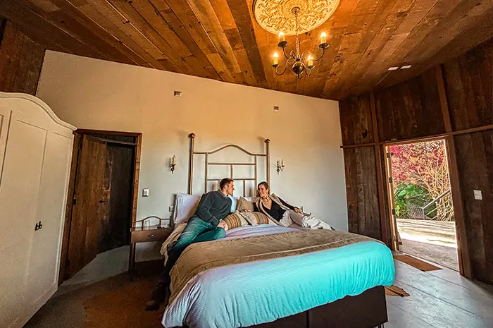 Dormitorio rústico en Hotel Santa Cruz con dos personas conversando sobre la cama, techo de madera, lámpara de araña, ventana con vista al jardín y ambiente cálido. La escena transmite intimidad pausada, estética territorial y conexión con Cava Colchagua.