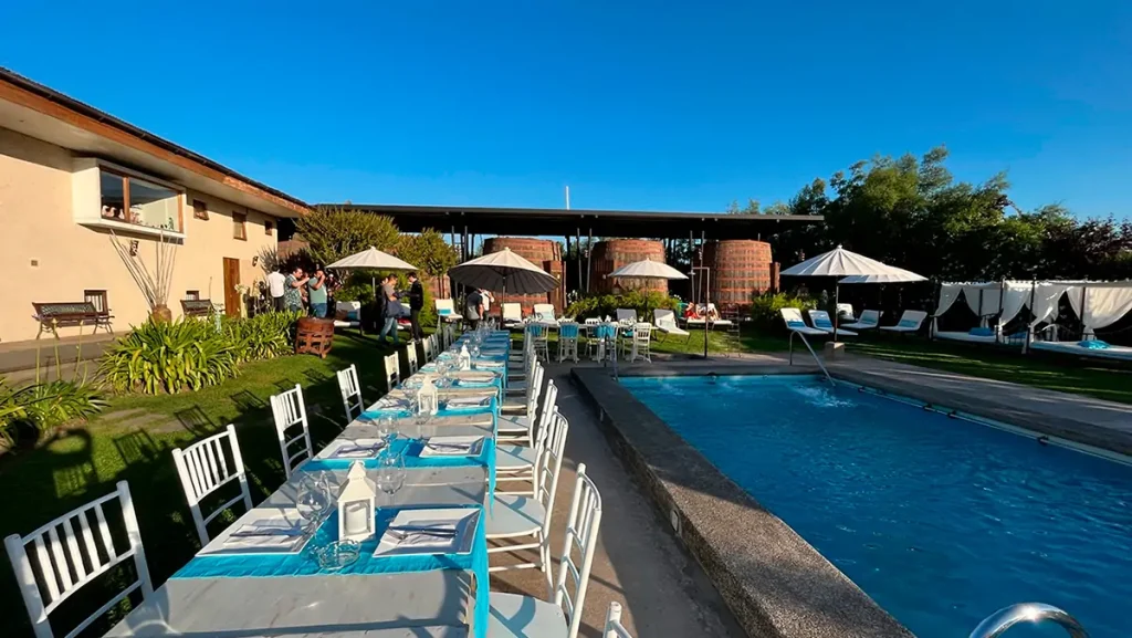 Evento junto a piscina en Hotel Santa Cruz con mesas largas vestidas, sillas blancas, personas conversando y fondo con camas bajo toldos. Cielo despejado y ambiente festivo. La escena transmite celebración corporativa, estética solar y conexión con Cava Colchagua.