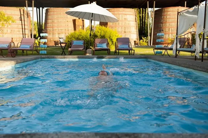 Piscina exterior en Hotel Santa Cruz con persona nadando, reposeras, sombrilla blanca y estructuras cilíndricas tipo barrica. Fondo con vegetación. La escena transmite relajo corporal, estética vinícola y conexión con Cava Colchagua.