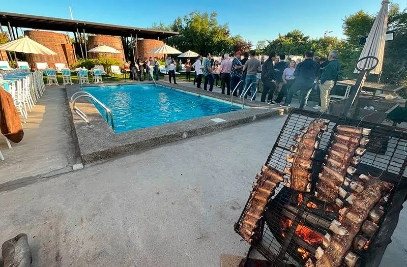 Evento exterior en Hotel Santa Cruz con piscina, grupo de personas conversando y parrilla con costillares sobre brasas. Reposeras, sombrillas blancas y ambiente festivo. La escena transmite relajo corporativo, estética gastronómica y conexión con Cava Colchagua.