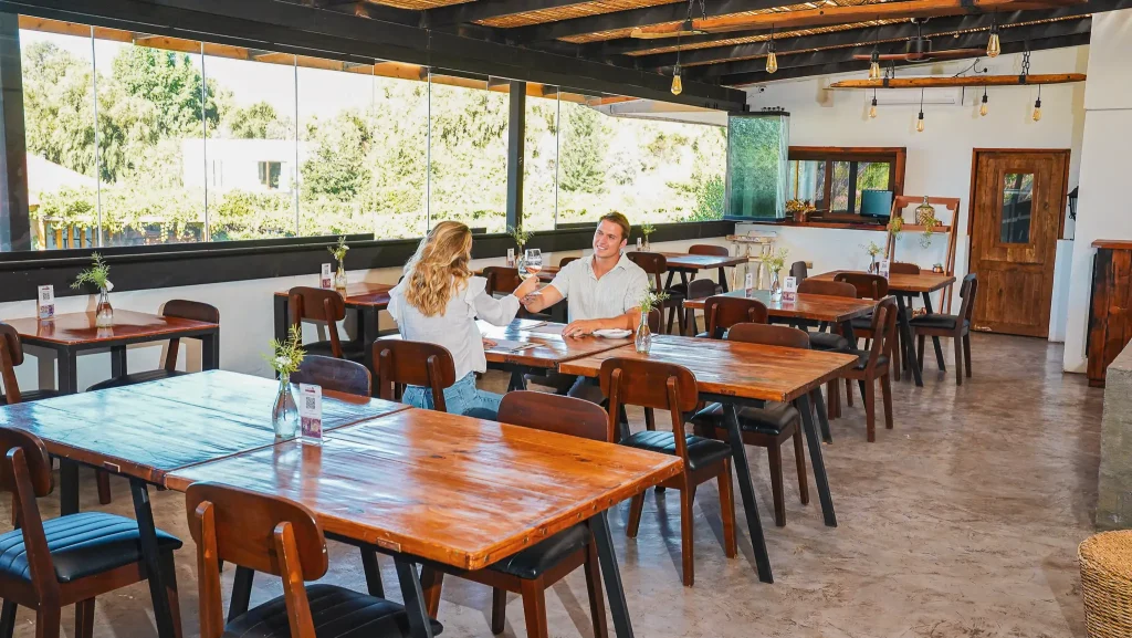 Restaurante interior en Hotel Santa Cruz con mesas y sillas de madera, floreros con plantas verdes, luz natural desde ventanales, techo con vigas expuestas y lámparas colgantes. Dos personas conversan en una mesa. La escena transmite calidez cotidiana, diseño rústico y conexión con Cava Colchagua.