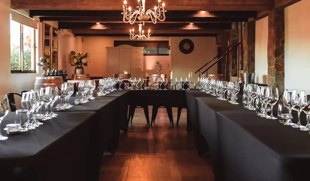 Salón de cata en Hotel Santa Cruz con mesas en forma de U, copas alineadas, vigas de madera, muros de piedra, flores y barricas decorativas. Ambiente rústico y elegante. La escena transmite ritual vinícola, estética institucional y conexión con Cava Colchagua.