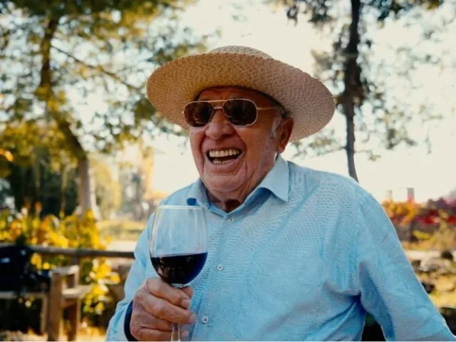 Don Raúl Ravanal, patriarca fundador del Hotel Cava Colchagua en Santa Cruz, sostiene una copa de vino de su viña que es parte de Hotel, él a trabajado este ensamblaje y sonríe a posando cerca de la laguna