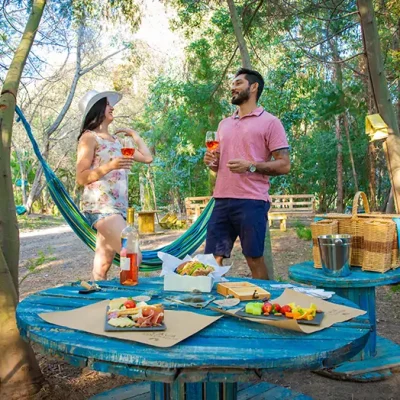 Picnic entre árboles en Hotel Santa Cruz con mesa azul rústica, comida variada, vino rosado y dos personas brindando. Hamaca al fondo, canasto y cubeta visibles. La escena transmite celebración silvestre, estética estacional y conexión con Cava Colchagua.